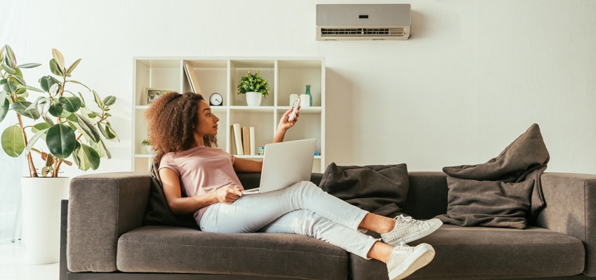 pretty-african-american-woman-using-laptop_web Woman sitting on couch using laptop and adjusting ductless mini split temperature with remote control.